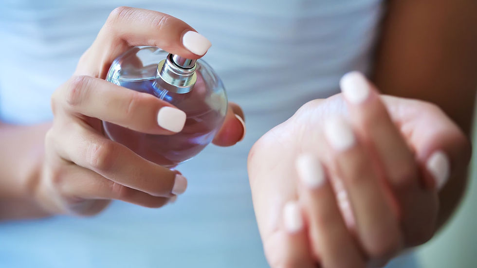 Close-up of a person spraying perfume on their wrist. The bottle is round and transparent, and nails are painted white. Soft, blurred background.
