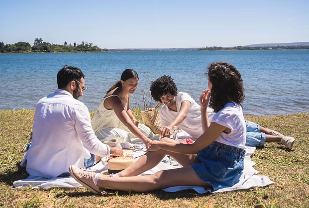 Four friends enjoy a picnic by a lake, sitting on a blanket. They're smiling, surrounded by food baskets. The sky is clear and sunny.