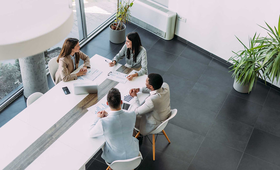 Four people in a modern office having a meeting around a white table with laptops and documents. Bright room, plants, and large windows.